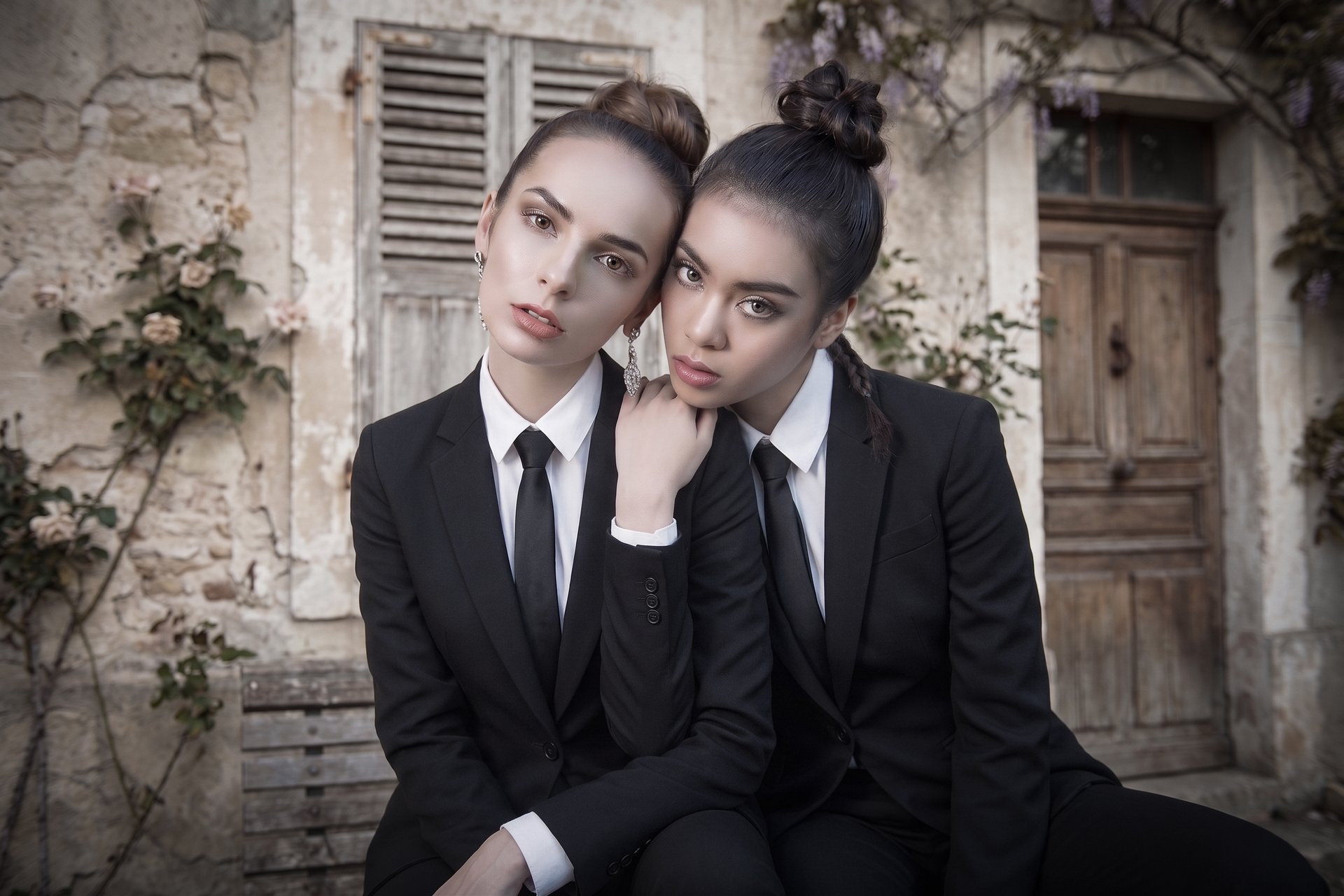 Two models in tailored black suits against European stone wall
